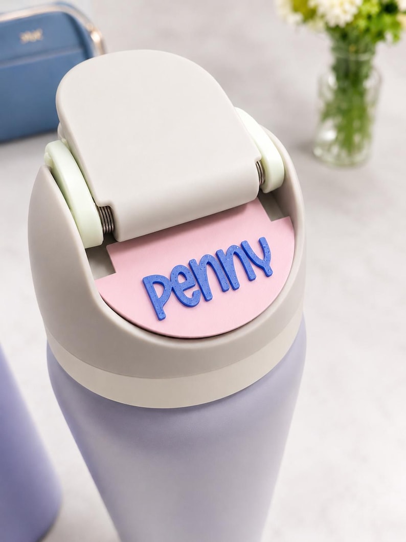 May include: A light purple water bottle with a beige lid featuring a hinged flip-top mechanism. A semi-circular pink name tag is attached to the front of the lid, displaying the name penny in raised, bold, dark blue lettering.