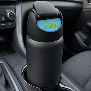 May include: A matte black insulated water bottle with a flip-top lid. The lid features a light blue rectangular insert with the name John written in a bright lime green cursive script. The bottle is cylindrical and fits into a vehicle cup holder.