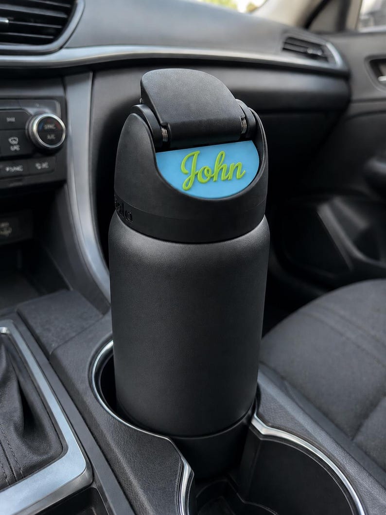 May include: A matte black insulated water bottle with a flip-top lid. The lid features a light blue rectangular insert with the name John written in a bright lime green cursive script. The bottle is cylindrical and fits into a vehicle cup holder.