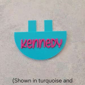 May include: A turquoise semi-circular plastic tag featuring the name Kennedy in raised magenta lettering. The tag has two rectangular tabs extending from the top edge. The text is written in a rounded, lowercase font style.