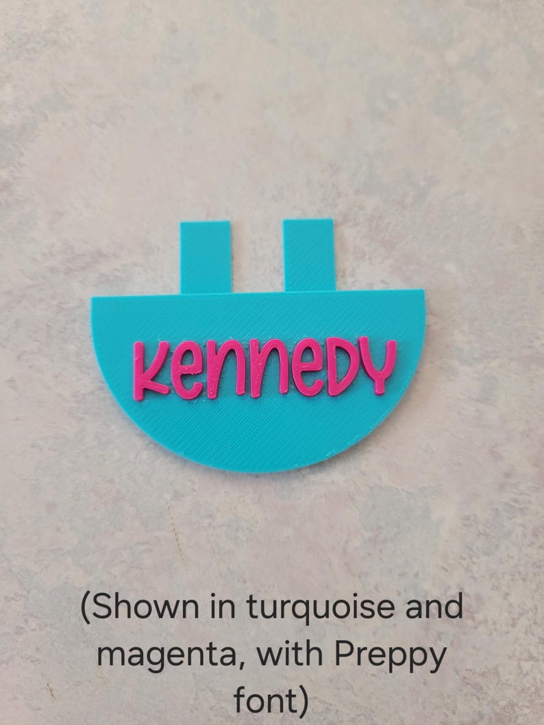 May include: A turquoise semi-circular plastic tag featuring the name Kennedy in raised magenta lettering. The tag has two rectangular tabs extending from the top edge. The text is written in a rounded, lowercase font style.