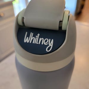 May include: A light blue water bottle featuring a gray flip-top lid with a dark blue circular insert. The name Whitney is printed in white script lettering on the dark blue insert. The bottle has a smooth matte finish and a light gray collar.