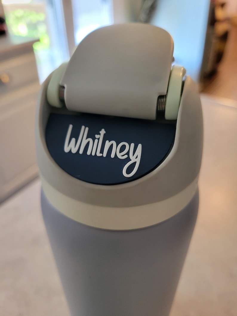 May include: A light blue water bottle featuring a gray flip-top lid with a dark blue circular insert. The name Whitney is printed in white script lettering on the dark blue insert. The bottle has a smooth matte finish and a light gray collar.