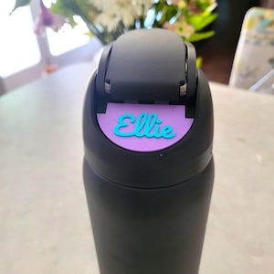 May include: A matte black water bottle featuring a black flip-top lid. A purple rectangular name tag is attached to the front of the lid, displaying the name Ellie in a raised, bright teal cursive script font. The bottle has a smooth finish.