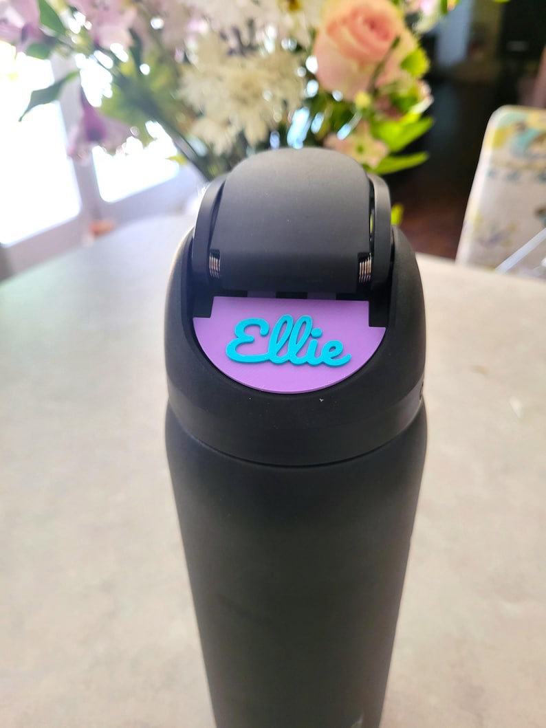 May include: A matte black water bottle featuring a black flip-top lid. A purple rectangular name tag is attached to the front of the lid, displaying the name Ellie in a raised, bright teal cursive script font. The bottle has a smooth finish.