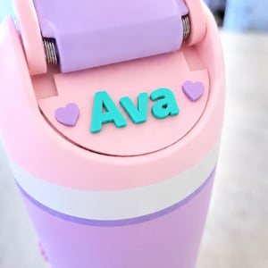 May include: A cylindrical water bottle featuring a light pink and lavender color scheme with a white horizontal band. The lid has a hinged lavender top and a flat section displaying the name Ava in teal letters, flanked by two small lavender hearts.