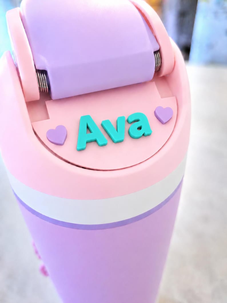 May include: A cylindrical water bottle featuring a light pink and lavender color scheme with a white horizontal band. The lid has a hinged lavender top and a flat section displaying the name Ava in teal letters, flanked by two small lavender hearts.