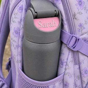 May include: A dark gray Owala water bottle with a pink name tag reading Sarah on the lid, placed in the side mesh pocket of a light purple backpack featuring a floral pattern with small purple flowers and thin dark stems throughout the fabric.