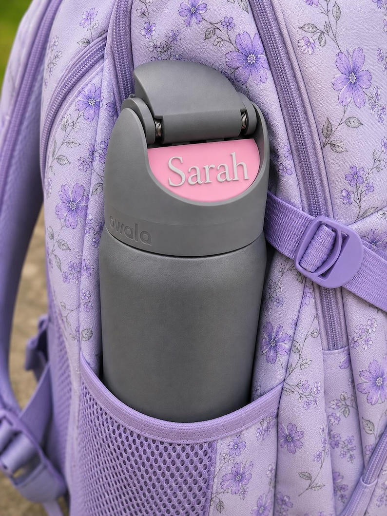 May include: A dark gray Owala water bottle with a pink name tag reading Sarah on the lid, placed in the side mesh pocket of a light purple backpack featuring a floral pattern with small purple flowers and thin dark stems throughout the fabric.