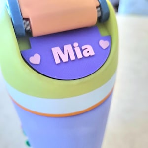 May include: A multi-colored water bottle featuring a lime green top section, a white middle band, and a lavender base. The lid has a peach-colored flip-top with blue hinges. A purple label on the lid displays the name Mia in pink with two hearts.