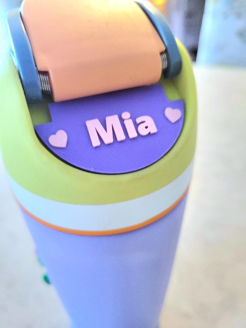 May include: A multi-colored water bottle featuring a lime green top section, a white middle band, and a lavender base. The lid has a peach-colored flip-top with blue hinges. A purple label on the lid displays the name Mia in pink with two hearts.