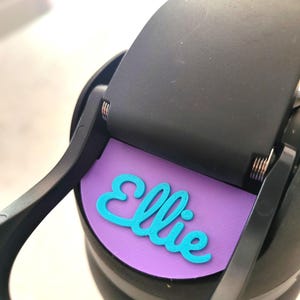 May include: A black plastic water bottle lid featuring a circular purple insert with the name Ellie written in raised, bright turquoise script lettering. The lid includes a hinged black cap mechanism with visible metal springs at the hinge point.