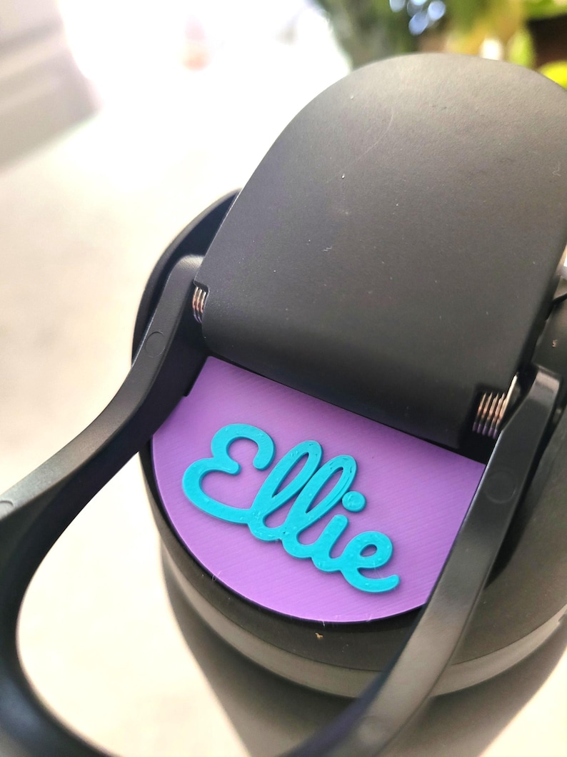 May include: A black plastic water bottle lid featuring a circular purple insert with the name Ellie written in raised, bright turquoise script lettering. The lid includes a hinged black cap mechanism with visible metal springs at the hinge point.