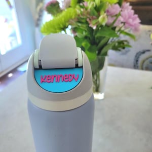 May include: A light purple water bottle with a gray flip-top lid featuring a bright blue accent plate. The name Kennedy is embossed in pink lettering on the blue plate. The bottle has a smooth, matte finish and a cylindrical shape.