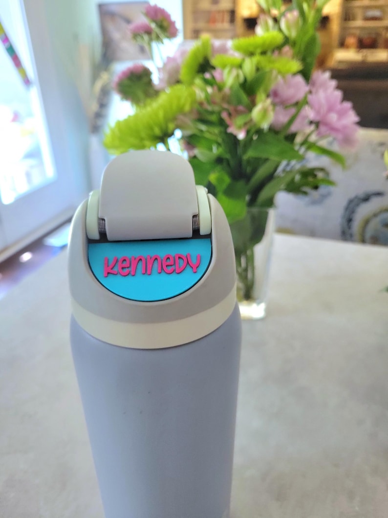 May include: A light purple water bottle with a gray flip-top lid featuring a bright blue accent plate. The name Kennedy is embossed in pink lettering on the blue plate. The bottle has a smooth, matte finish and a cylindrical shape.