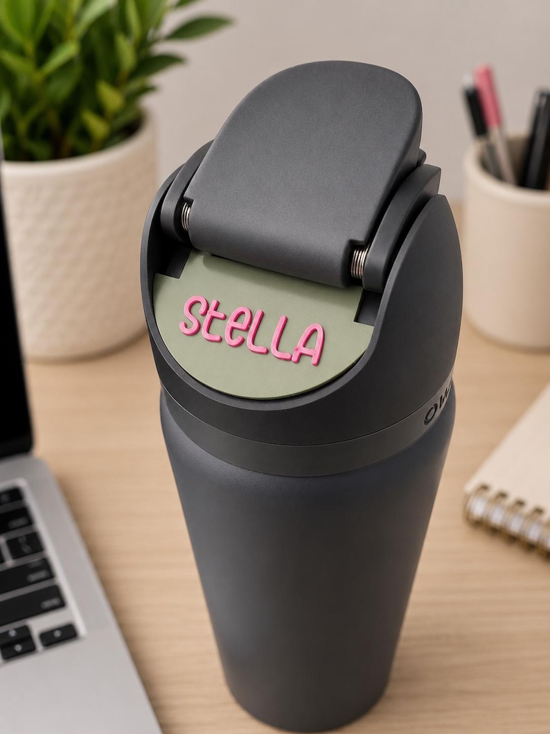 May include: A dark gray insulated tumbler with a hinged lid featuring a sage green circular insert. The name Stella is embossed in pink lettering on the green insert. The tumbler has a tapered body and a matte finish, showing a functional design.