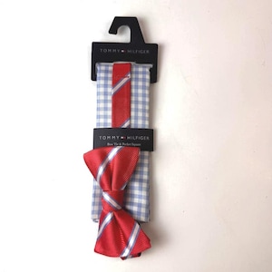 May include: A Tommy Hilfiger bow tie and pocket square set. The bow tie is red with diagonal white and blue stripes. The pocket square features a light blue and white gingham check pattern. The set is attached to a black branded display card.