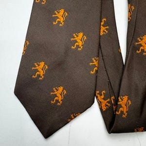 May include: A dark brown necktie featuring a repeating pattern of small, orange, rampant lion figures. The fabric has a visible diagonal twill weave texture throughout the entire surface of the tie, which is displayed in a flat, unfolded position.