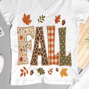 May include: A white V-neck t-shirt featuring the word FALL in large letters. Each letter has a different pattern: floral, leopard print, orange gingham, and olive green with white dots. The design includes scattered autumn leaves and acorns.