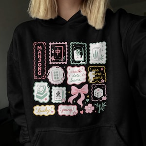 Könnte beinhalten: Schwarzer Kapuzenpullover mit einem farbenfrohen Mahjong-Motiv. Das Design zeigt Spielsteine und Schriftzüge in Rosa, Grün und Weiß, darunter das Wort "Mahjong" und die Phrase "cracks dots bams".