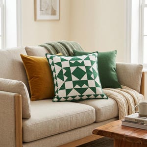 May include: A beige sofa with several decorative pillows. The pillows include a mustard yellow velvet pillow, a green and white geometric patterned pillow, and a solid green pillow. A green blanket and a cream-colored knitted throw are also on the sofa.