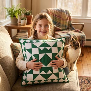 May include: A decorative square pillow with a green and white geometric pattern. The pillow is held by a person sitting on a gray sofa. A cat sits next to the person. Plants and a wooden table are in the background.