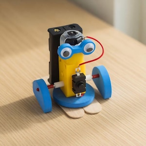 Op de afbeelding: Een kleine, handgemaakte robotspeelgoed met een gele behuizing, blauwe wielen en wiebelogen. De robot heeft een zwart batterijpakket en een rode draad die de componenten verbindt. Het is gebouwd op een basis van houten knutselstokjes.