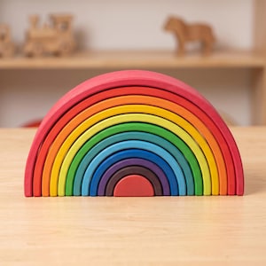 Pode incluir: Um brinquedo de empilhar arco-íris de madeira com uma variedade vibrante de cores, incluindo vermelho, laranja, amarelo, verde, azul e roxo. O brinquedo é feito de várias peças arqueadas que se encaixam. Está sobre uma superfície de madeira clara.