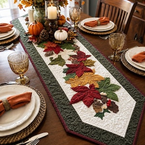 May include: A quilted table runner featuring a central white panel decorated with appliqued fabric leaves in shades of red, orange, yellow, and green, bordered by a dark green quilted fabric with a swirling pattern and a thin maroon edge.