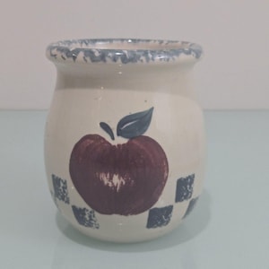 Może przedstawiać: Ceramiczny słoik w kremowym kolorze z ręcznie malowanym wzorem czerwonego jabłka i zielonym liściem. Obrzeże ma niebieski wzór w kratkę. Słoik ma około 10 cm wysokości.