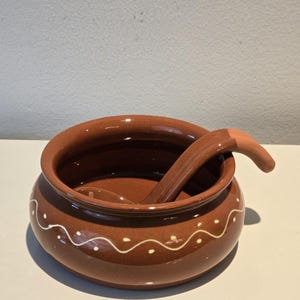 May include: A round, brown ceramic bowl with a glossy finish and a matching spoon resting inside. The exterior of the bowl features a hand-painted white wavy line pattern with small dots. The spoon has a curved handle that hooks over the rim.
