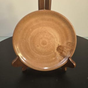 May include: A round, brown ceramic plate with a textured surface, displayed on a wooden stand. The plate has a slightly raised rim and a darker brown area on the right side. The wooden stand has a curved design.