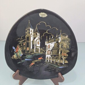 Può includere: Piatto decorativo nero con un'illustrazione dorata e bianca di una scena cittadina europea. Il piatto ha una forma unica ed è esposto su un supporto di legno. Il piatto ha la scritta "Longwy".