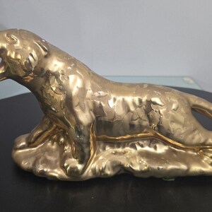 Könnte beinhalten: Goldfarbene Keramikfigur eines brüllenden Löwen. Der Löwe liegt in einer liegenden Position, mit offenem Maul und sichtbaren Zähnen. Die Figur hat eine strukturierte Oberfläche und steht auf einem Sockel.