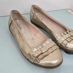 May include: A pair of gold-colored Clarks ballet flats. The shoes have a rounded toe and a decorative ruffle detail on the vamp. The insole features the Clarks logo and a floral design. The shoes are on a light blue surface.