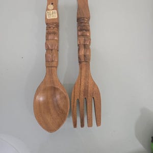 May include: A wooden spoon and fork set. The spoon has a rounded bowl, and the fork has four tines. Both utensils have carved handles with a decorative pattern. The wood has a warm, natural tone.