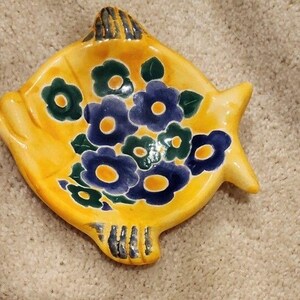 May include: A yellow ceramic fish-shaped dish with a floral design. The flowers are blue and green with yellow centers. The dish has a textured surface and is likely handmade.