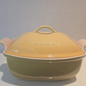 May include: A yellow Le Creuset Dutch oven with a matching lid. The lid has a loop handle and the brand name embossed on the top. The oven has a light green base with pink handles.