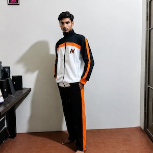 Puede incluir: Un chándal negro, blanco y naranja compuesto por una chaqueta con cremallera y un pantalón a juego. La chaqueta presenta un torso blanco, mangas negras con rayas naranjas y un cuello negro. El pantalón negro tiene una raya naranja vertical en el lateral.