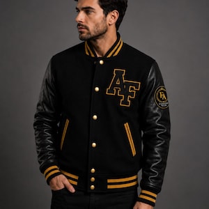 Könnte beinhalten: Schwarze Collegejacke mit goldenen Akzenten, einschließlich Lederärmeln und Knopfverschluss. Die Jacke hat goldene Streifen an Kragen, Manschetten und Saum, mit den Buchstaben "AF" und einem runden Aufnäher auf der Brust.