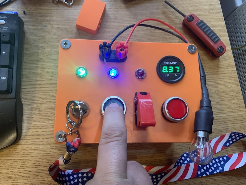 Może przedstawiać: Pomarańczowy elektroniczny panel sterowania z cyfrowym wyświetlaczem napięcia pokazującym 8,37 DC. Panel posiada r&oacute;żne przyciski, przełączniki i wskaźniki, w tym zielone i niebieskie diody LED. Palec naciska przycisk.
