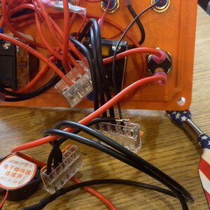 Może przedstawiać: Zbliżenie pomarańczowej skrzynki z komponentami elektrycznymi z r&oacute;żnymi przewodami, złączami i małym głośnikiem. Czerwone i czarne przewody są podłączone do skrzynki, wraz z małym metalowym narzędziem z patriotycznym amerykańskim wzorem flagi.