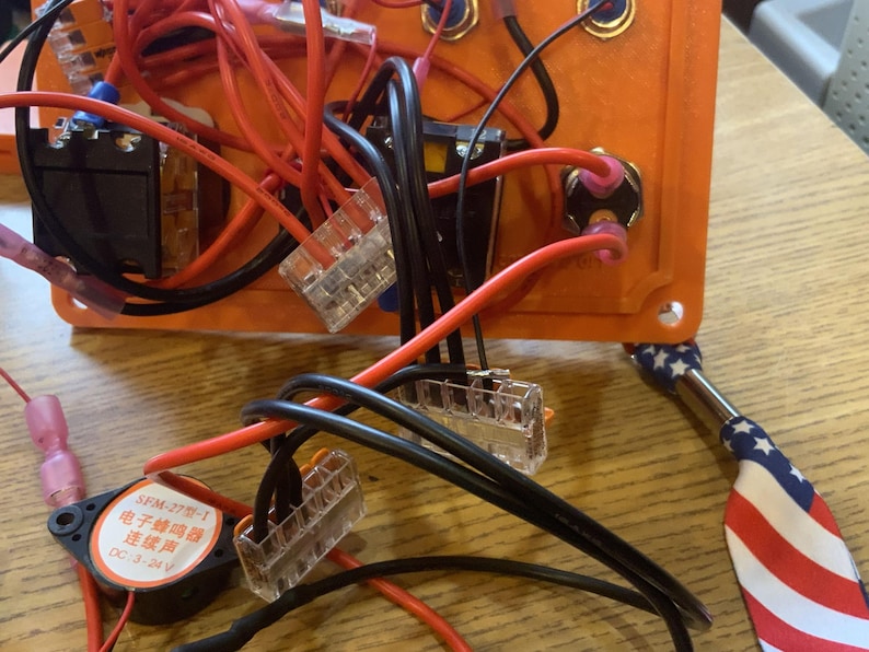 Może przedstawiać: Zbliżenie pomarańczowej skrzynki z komponentami elektrycznymi z r&oacute;żnymi przewodami, złączami i małym głośnikiem. Czerwone i czarne przewody są podłączone do skrzynki, wraz z małym metalowym narzędziem z patriotycznym amerykańskim wzorem flagi.