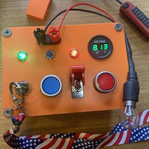 Może przedstawiać: Pomarańczowy panel sterowania elektronicznego z cyfrowym wyświetlaczem napięcia wskazującym 8,13 V DC. Zawiera zieloną diodę LED, niebieskie i czerwone przyciski, czerwony przełącznik i żar&oacute;wkę. Zawiera brelok i czerwono-czarny kabel.