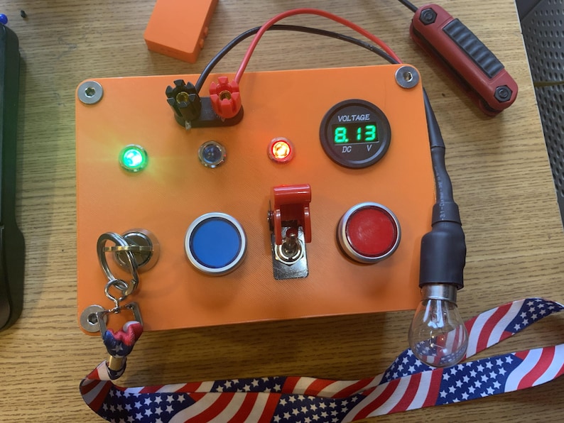 Może przedstawiać: Pomarańczowy panel sterowania elektronicznego z cyfrowym wyświetlaczem napięcia wskazującym 8,13 V DC. Zawiera zieloną diodę LED, niebieskie i czerwone przyciski, czerwony przełącznik i żar&oacute;wkę. Zawiera brelok i czerwono-czarny kabel.