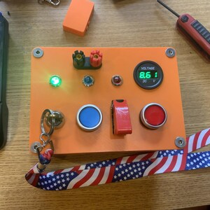 Może przedstawiać: Pomarańczowy elektroniczny panel sterowania z r&oacute;żnymi komponentami, w tym zieloną diodą LED, cyfrowym wyświetlaczem napięcia pokazującym 8,6 V oraz czerwonymi i niebieskimi przyciskami. Dołączona jest smycz z amerykańskim wzorem flagi. Widoczne są r&oacute;wnież czerwone narzędzie i klawiatura.