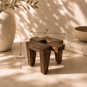 Könnte beinhalten: Ein dunkelbrauner dekorativer Holzständer, 10,2 cm hoch und 12,7 cm breit, mit quadratischer Oberseite und vier Beinen. Der Ständer hat eine natürliche Holzmaserung und ein minimalistisches Design. Der Ständer steht neben einem Buch mit dem Titel "KINFOLK".