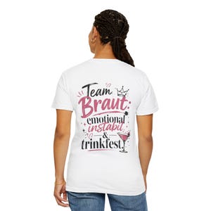 Könnte beinhalten: Ein weißes T-Shirt mit kurzen Ärmeln und einem Aufdruck auf der Rückseite mit dem Text Team Braut: emotional instabil & trinkfest. Das Design enthält eine kleine Krone, Sterne, Herzen und eine Illustration eines rosa Cocktails in Schwarz und Pink.
