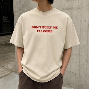 May include: Beige t-shirt with the text "DON'T BULLY ME I'LL COME" in red. The shirt has short sleeves and a crew neck. The model is wearing brown pants and has their hands in their pockets.