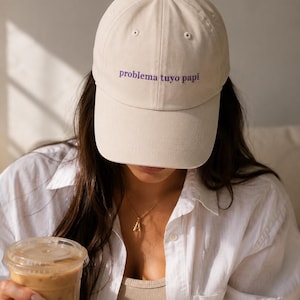 May include: A cream-coloured baseball cap with the phrase "problema tuyo papi" embroidered in purple. The cap is worn by a person wearing a white button-down shirt and holding a beverage in a clear plastic cup.